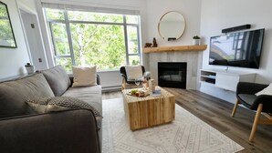 A modern living room with a sofa, coffee table, fireplace, TV, and large windows. - 2BR 2BA Village Escape w/ Updated Hot Tub, Perfect Location & Mountain Views (Whistler)