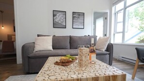 A modern living room with a grey sofa, a wooden coffee table with a bottle of wine and glasses, and two framed pictures on the wall. - 2BR 2BA Village Escape w/ Updated Hot Tub, Perfect Location & Mountain Views (Whistler)