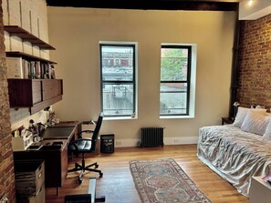 A bedroom with a bed, a desk with a chair, a radiator, and two windows.
