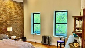 Un dormitorio con pared de ladrillo, dos ventanas con vistas a la vegetación, un radiador, una cama, un escritorio con silla y una pequeña mesa con un cuenco.