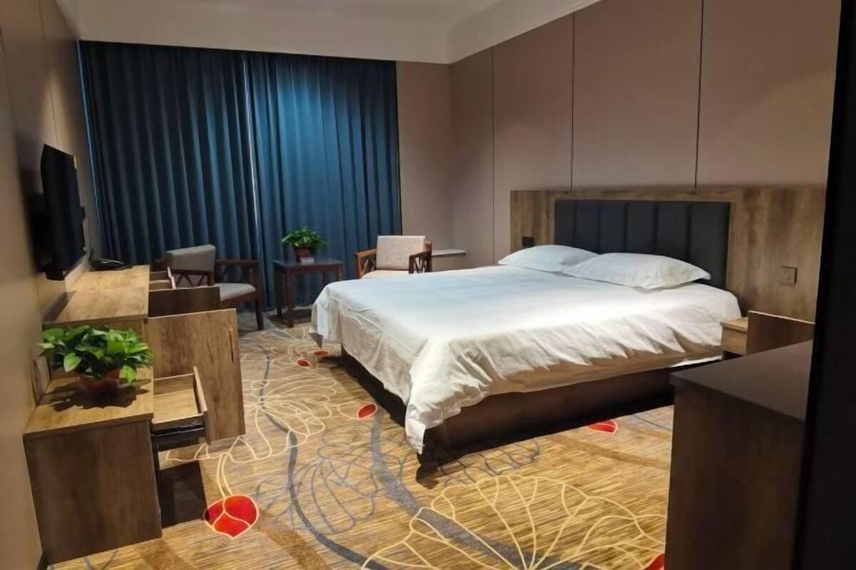 A modern hotel room with a large bed, a TV mounted on the wall, a seating area with chairs and a small table, and a decorative carpet.