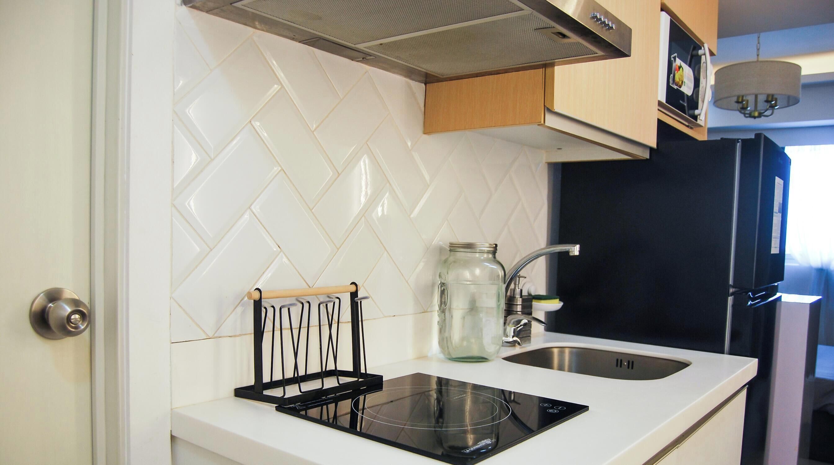 Una cocina con baldosas blancas tipo espiga, una placa de inducción negra, un lavamanos de acero inoxidable y armarios de madera.