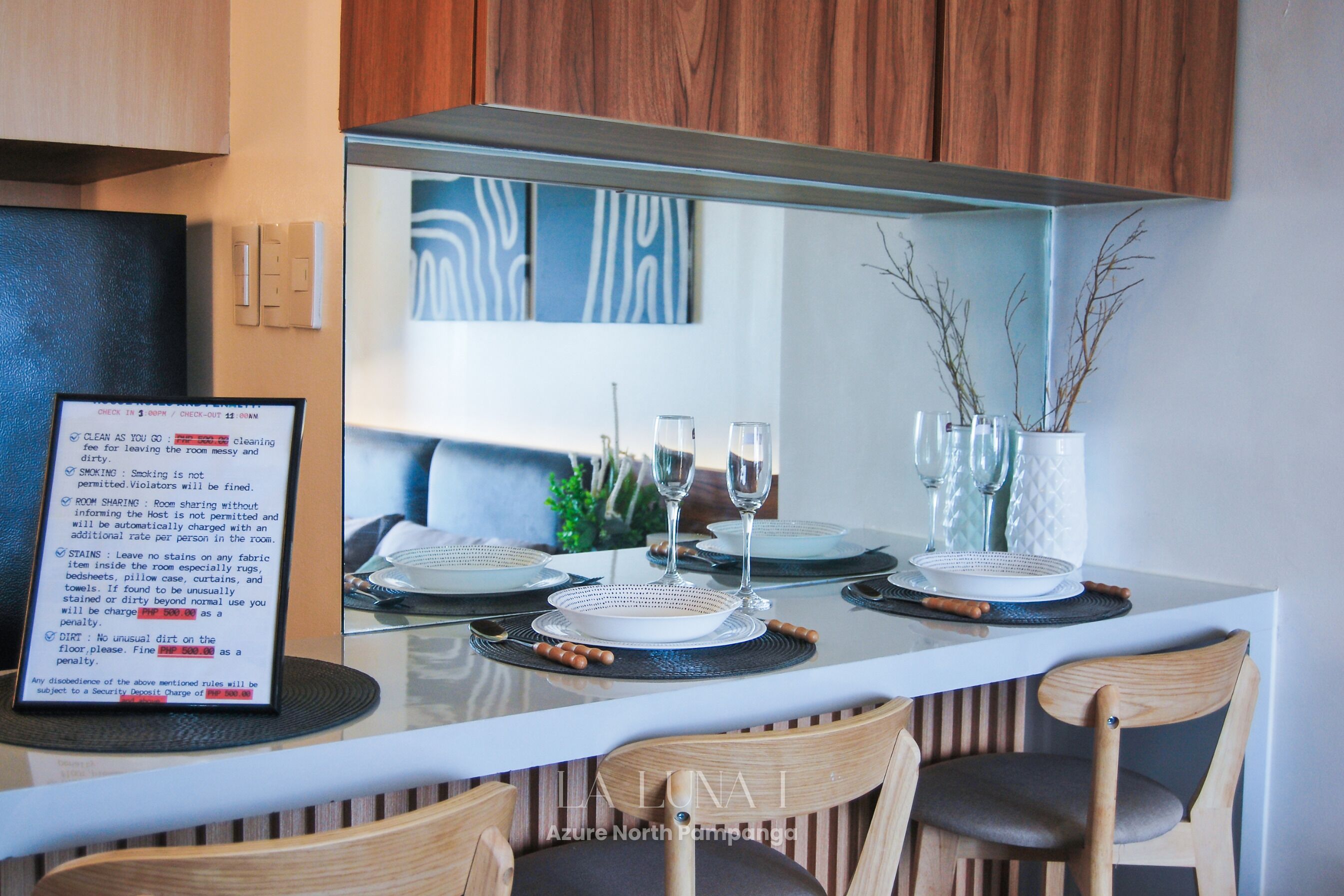 Un comedor moderno con una mesa de vidrio, platos blancos y dos copas de vino. Hay sillas de madera y un jarrón con ramas sobre la mesa.
