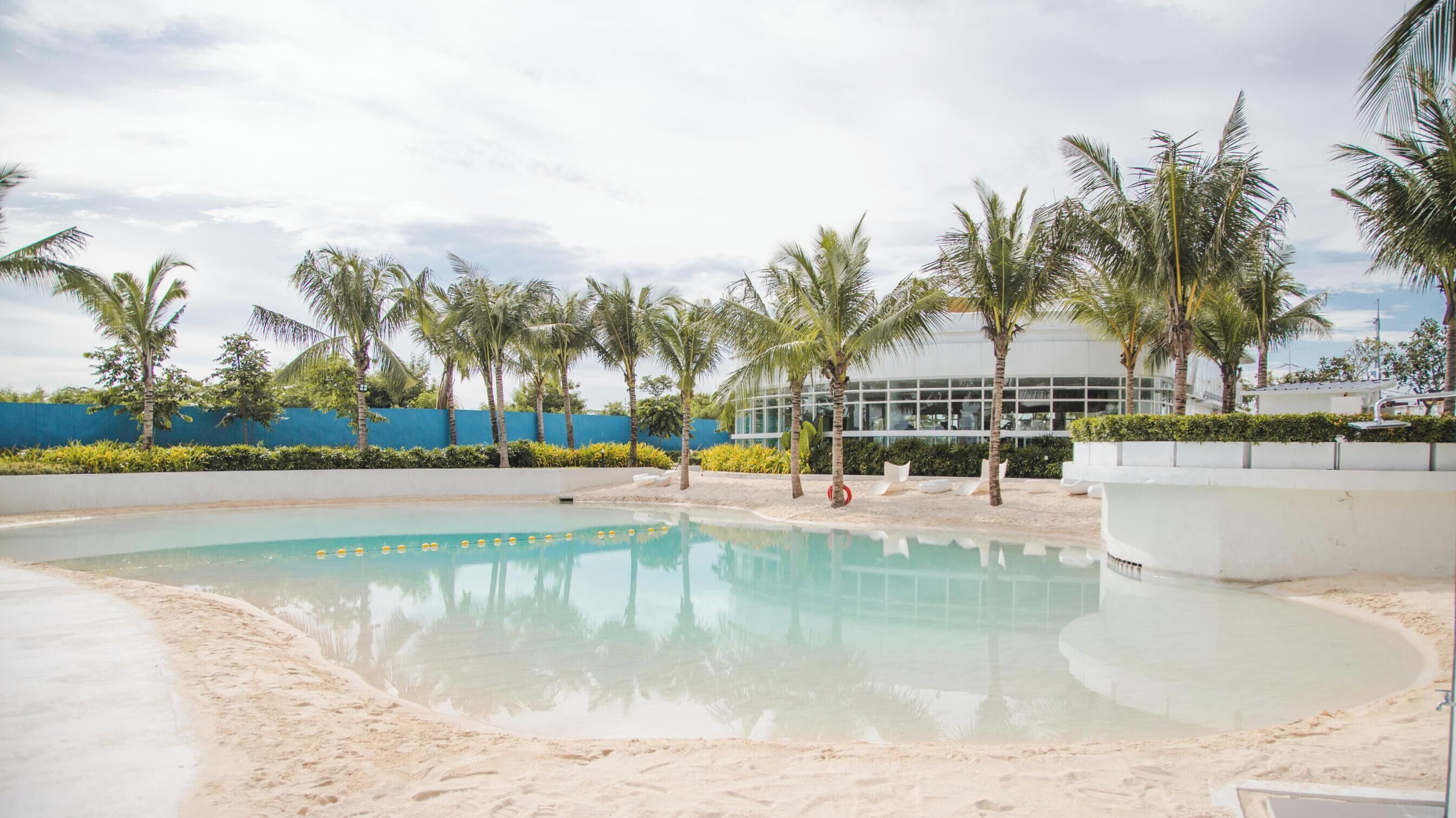 Ein Schwimmbecken mit Sandstrand, umgeben von Palmen und einem modernen Gebäude mit großen Fenstern.