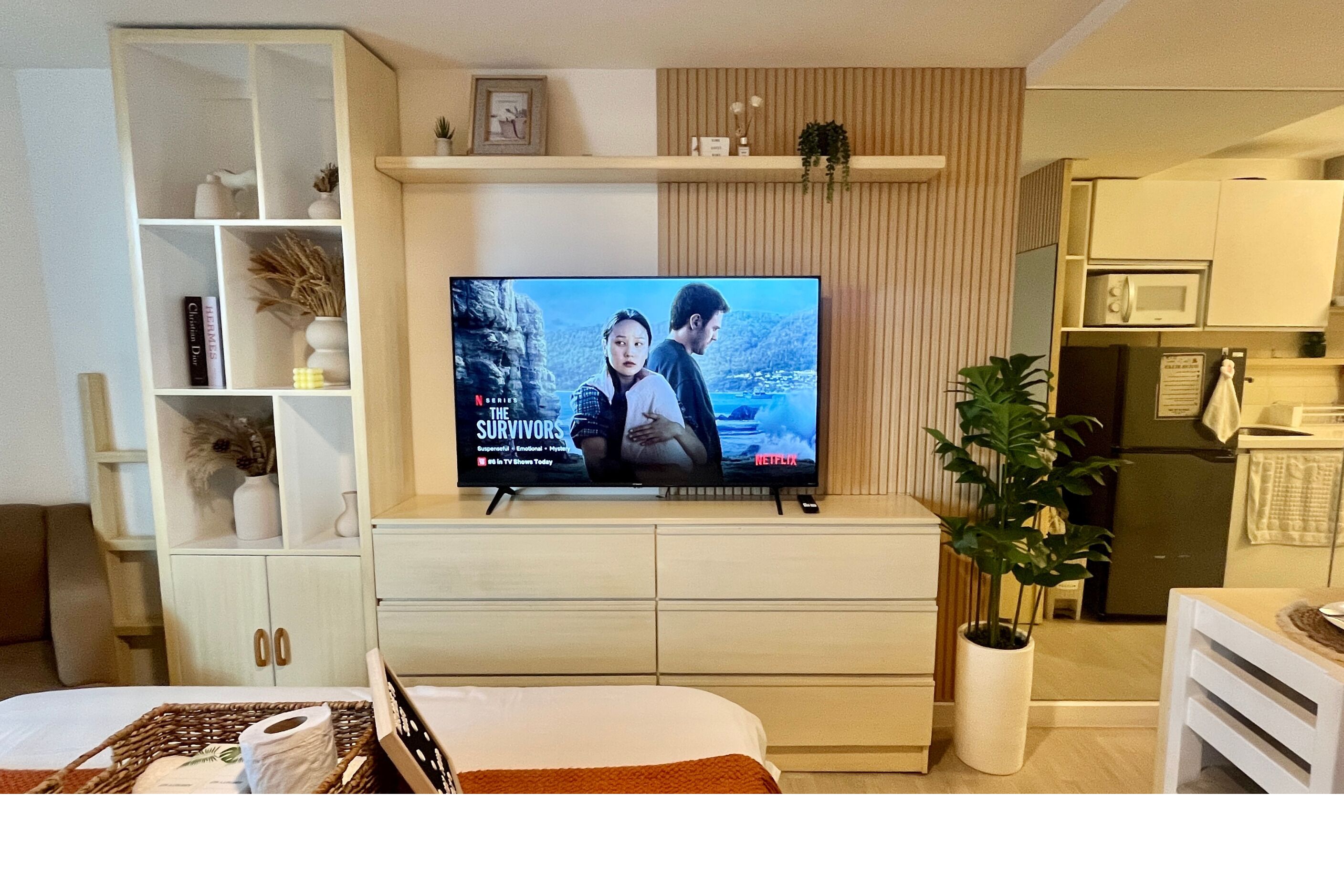 Un salon moderne avec un téléviseur à écran plat diffusant la série « The Survivors » sur Netflix, un meuble blanc avec des étagères, une plante en pot et un coin cuisine équipé d’un four à micro-ondes.