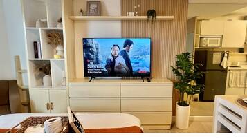 Un salon moderne avec un téléviseur à écran plat diffusant la série « The Survivors » sur Netflix, un meuble blanc avec des étagères, une plante en pot et un coin cuisine équipé d’un four à micro-ondes.