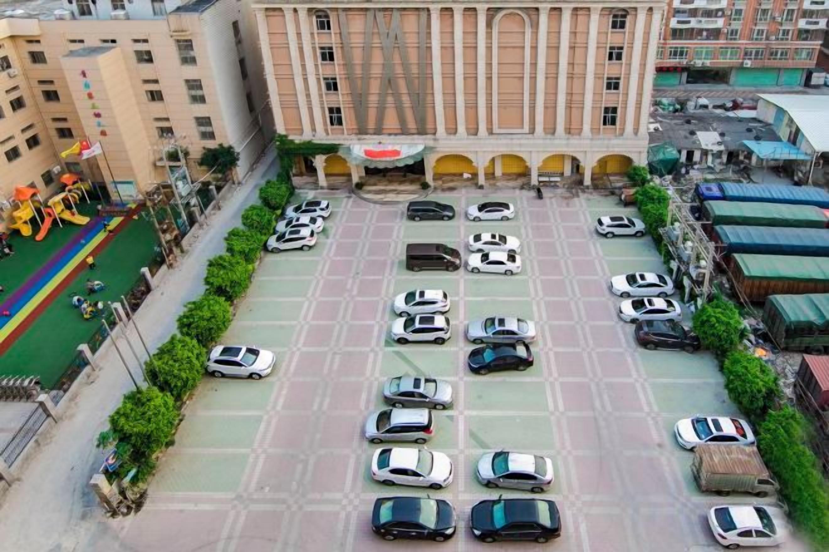 Un estacionamiento con varios automóviles, un edificio con columnas y un área verde con gente.