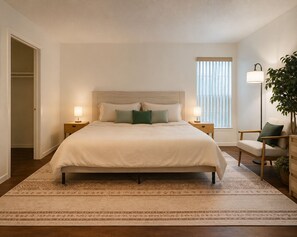 A bedroom with a large bed, bedside lamps, a chair, a floor lamp, and a window with blinds. - Living at the Palms (Tarpon Springs)