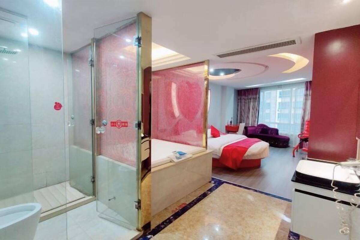 Une chambre d’hôtel dotée d’une douche en verre, d’un lit avec une literie rouge et d’une grande fenêtre.