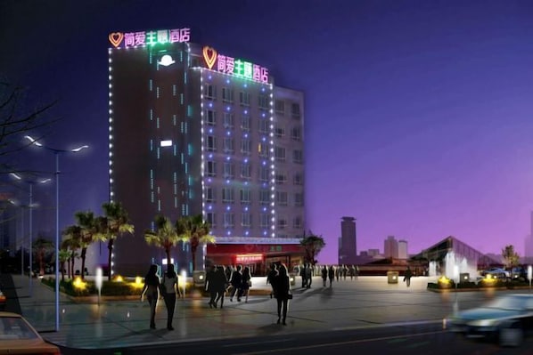 Um hotel com vários andares, fachada iluminada, área de pedestres com palmeiras e estacionamento.