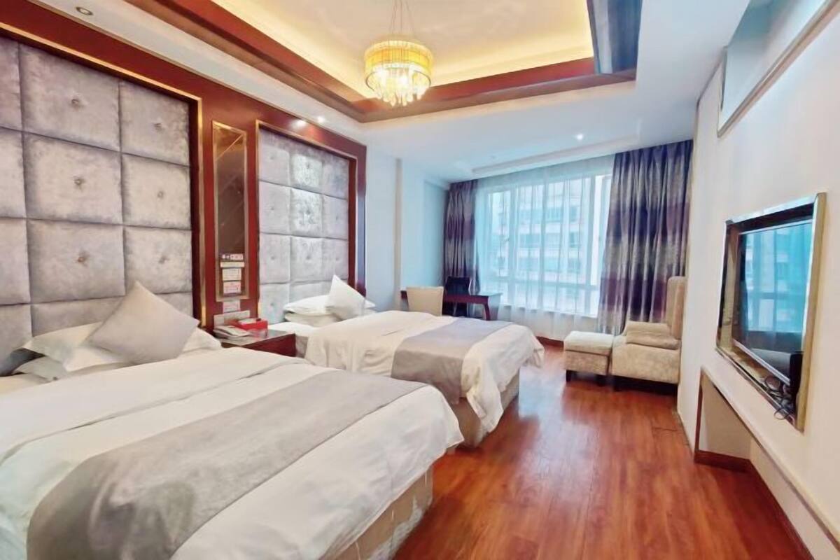 Une chambre d’hôtel avec deux lits, une télévision, un lustre et une grande fenêtre avec des rideaux.