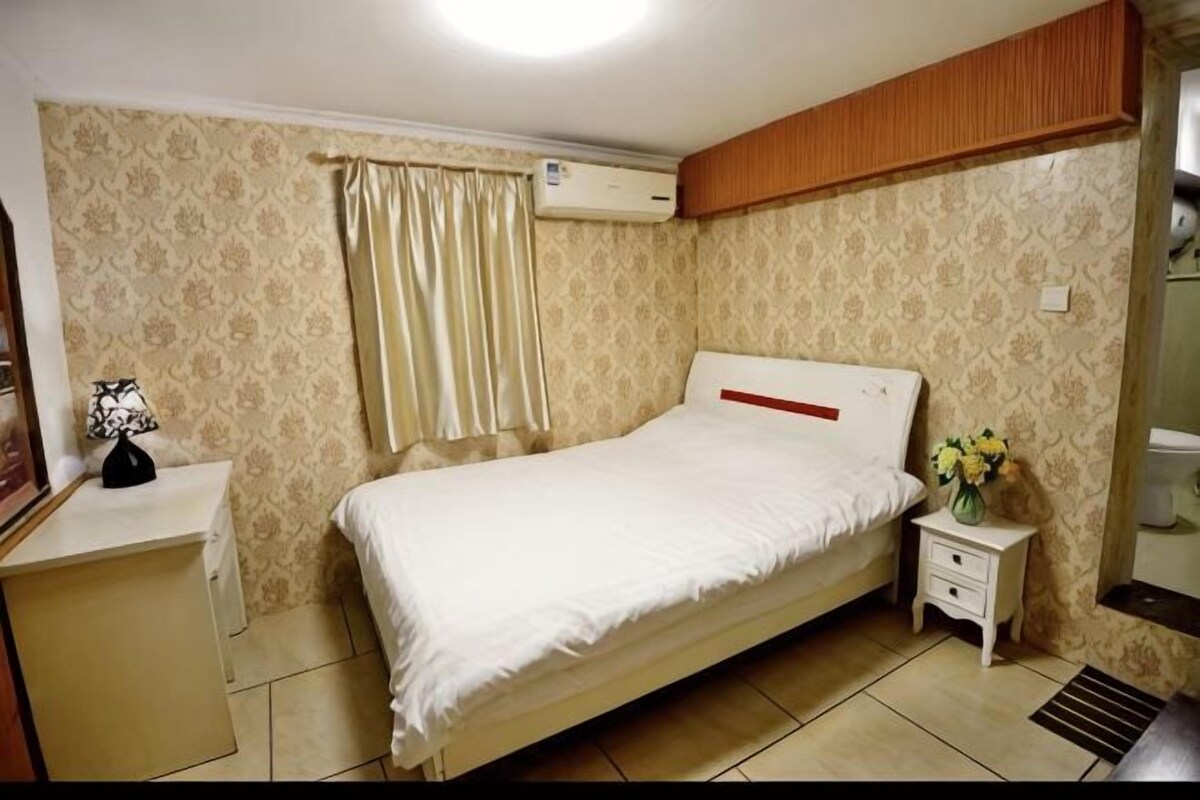 A bedroom with a single bed, a bedside table, a nightstand, an air conditioning unit, and a window with curtains.