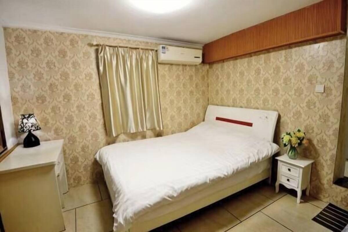 A bedroom with a bed, a nightstand, an air conditioning unit, and a window with curtains.
