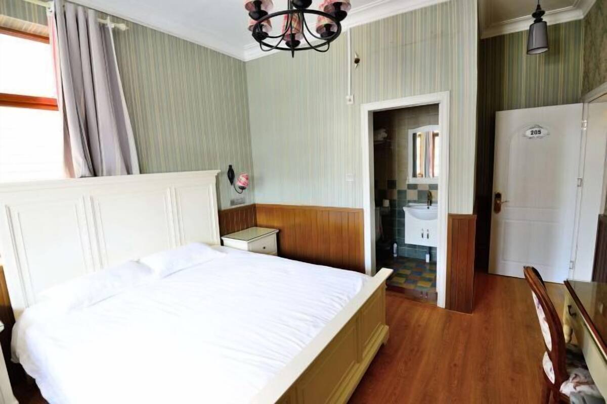 A bedroom with a bed, a nightstand, a wooden headboard, a chandelier, a bathroom with a sink, and a door with a number.