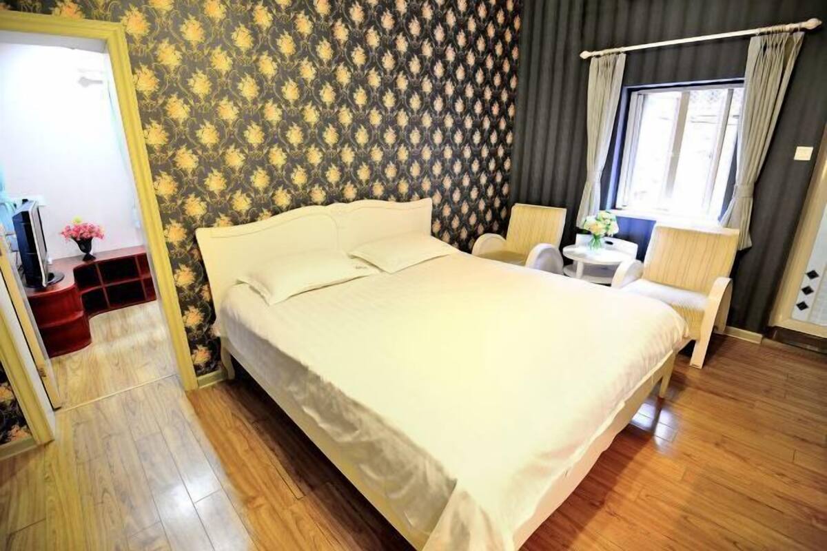 A hotel room with a bed, a small table with a chair, a window with curtains, and a patterned wallpaper.