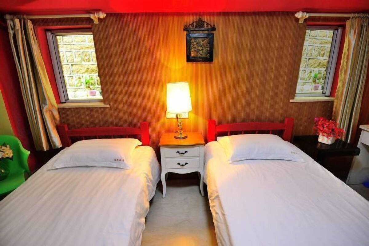 A room with two beds, a bedside table with a lamp, and a window with curtains.