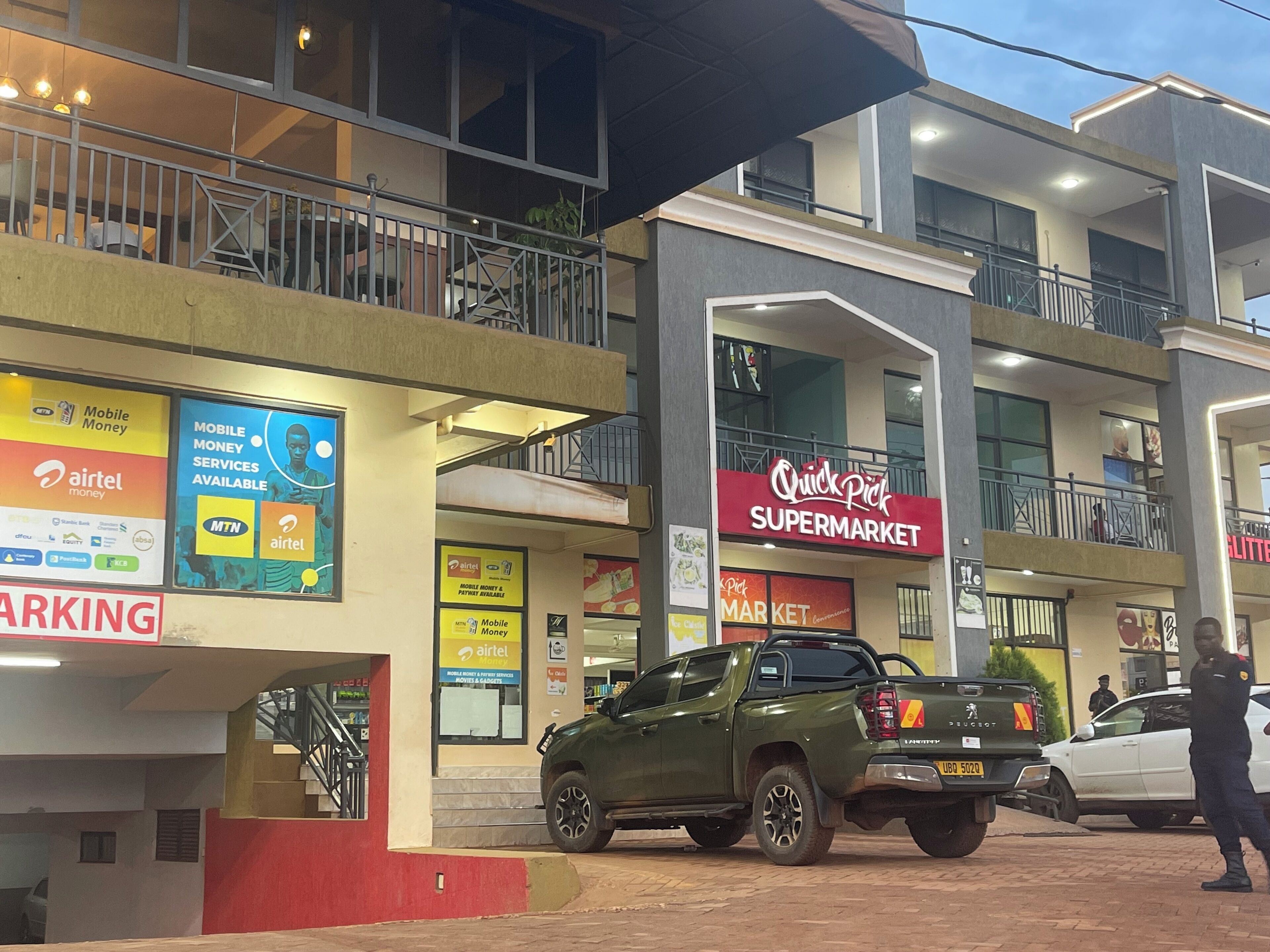 Um edifício de vários andares com um letreiro 'Quick Pick Supermarket', uma caminhonete verde estacionada em frente e um anúncio de 'Mobile Money' na fachada.