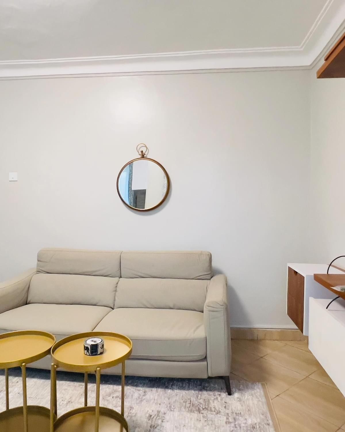 Ein Wohnzimmer mit einem beigefarbenen Sofa, zwei kleinen runden Tischen, einem runden Spiegel an der Wand und einem weißen Schrank.