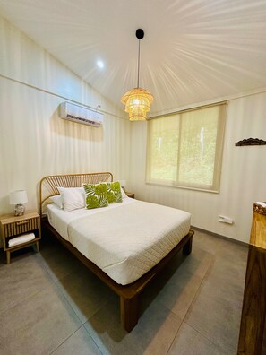 A bedroom with a bed, bedside table, lamp, air conditioning unit, window, and a hanging light fixture.