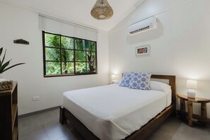 Un dormitorio con un ventanal grande, una cama con ropa de cama blanca, una mesita de noche con una lámpara y un equipo de aire acondicionado en la pared.