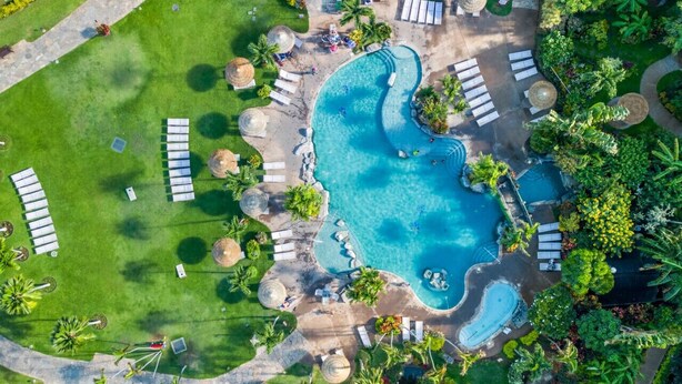 An aerial view of a resort with a large central pool, multiple lounge areas with umbrellas, and lush landscaping. - Spacious 2 Bed with Views! (Lahaina)