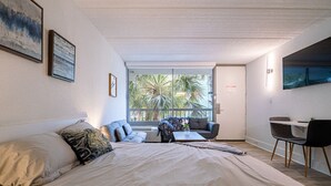 A bedroom with a large bed, a sofa, a small table, and a chair. There is a sliding glass door leading to a balcony with a view of palm trees.