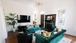 A living room with a teal sofa set, a black armchair, a coffee table, and a flat-screen TV. There is a fireplace with a green mantle and a large potted plant.