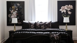 A bedroom with a black bed, two nightstands, a window with white curtains, and two floral paintings on the walls.