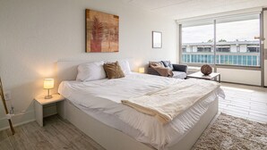A modern bedroom with a large bed, a bedside table with a lamp, a view of the outside through a large window, and a painting on the wall. - Lovely 1BR Suite Next to Disney With Lake view (Kissimmee)