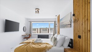 A modern hotel room with a large bed, a wooden headboard, a TV mounted on the wall, and a window with a view of buildings. - Seaside Escape: Warm & Cosy Winter Getaway by the Sea (Seaford)