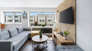 A modern living room with a grey sectional sofa, a round wooden coffee table, and a TV mounted on a geometric-patterned wall. - Seaside Escape: Warm & Cosy Winter Getaway by the Sea (Seaford)