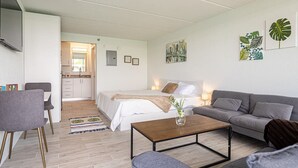 Un salon moderne et bien éclairé, avec un canapé, une table basse et un lit. On aperçoit une cuisine au fond de la pièce.