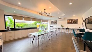 A modern dining area with a long table and clear chairs, large windows with a view of greenery, and a contemporary ceiling light fixture. - Stardust Boutique Hotel (Hua Hin)