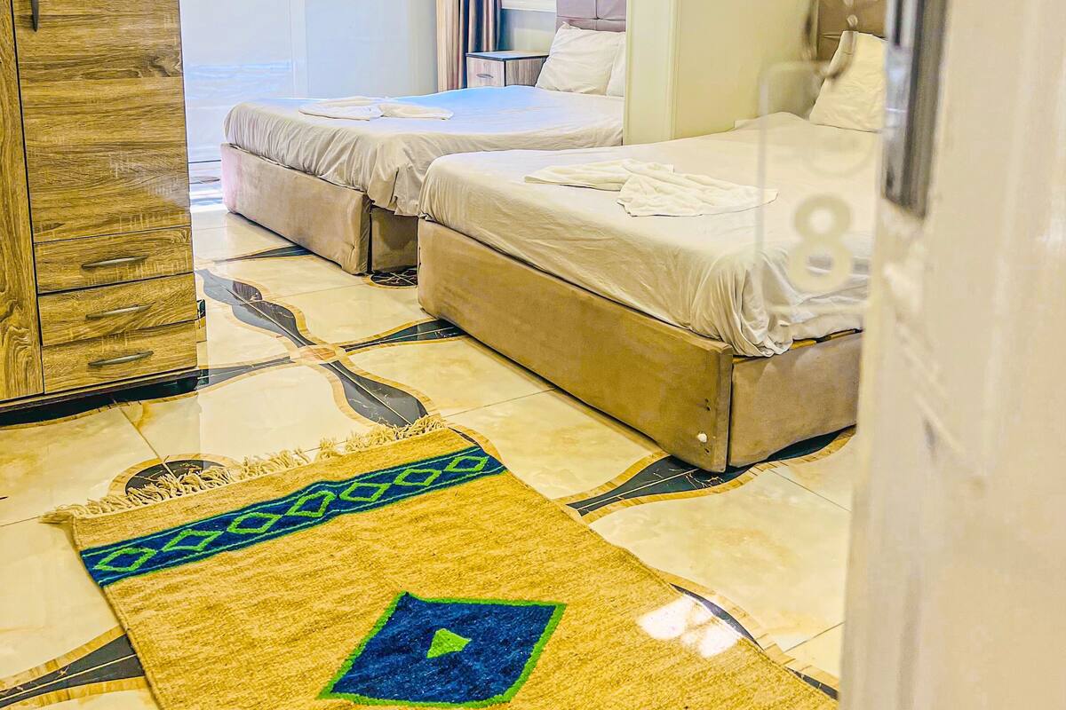 Habitación de hotel con dos camas, un armario de madera, una alfombra estampada y un baño visible a través de una puerta abierta.