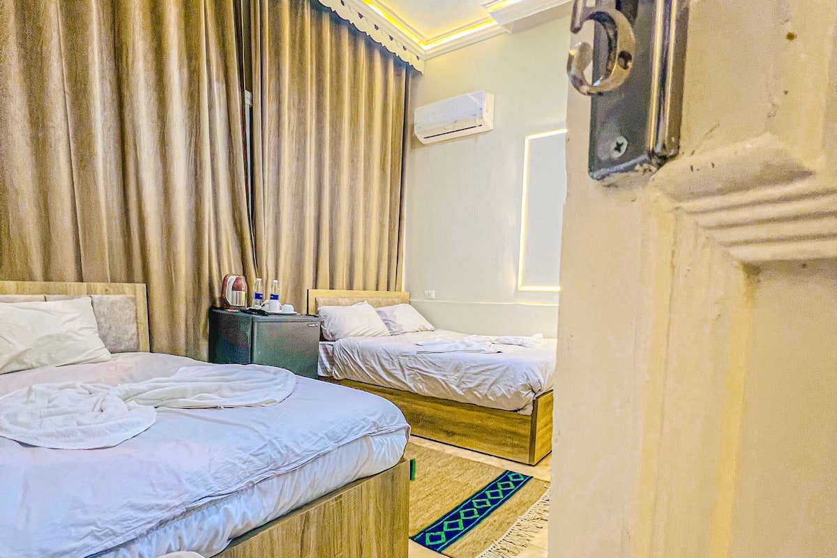 Habitación de hotel con dos camas, cabecero de madera, una mesita con lámpara y una alfombra azul estampada.