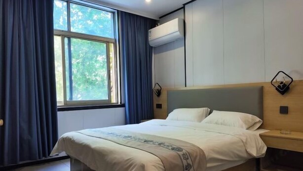 Una habitación de hotel con cama, ventana con cortinas, aire acondicionado y mesita de noche con lámpara.