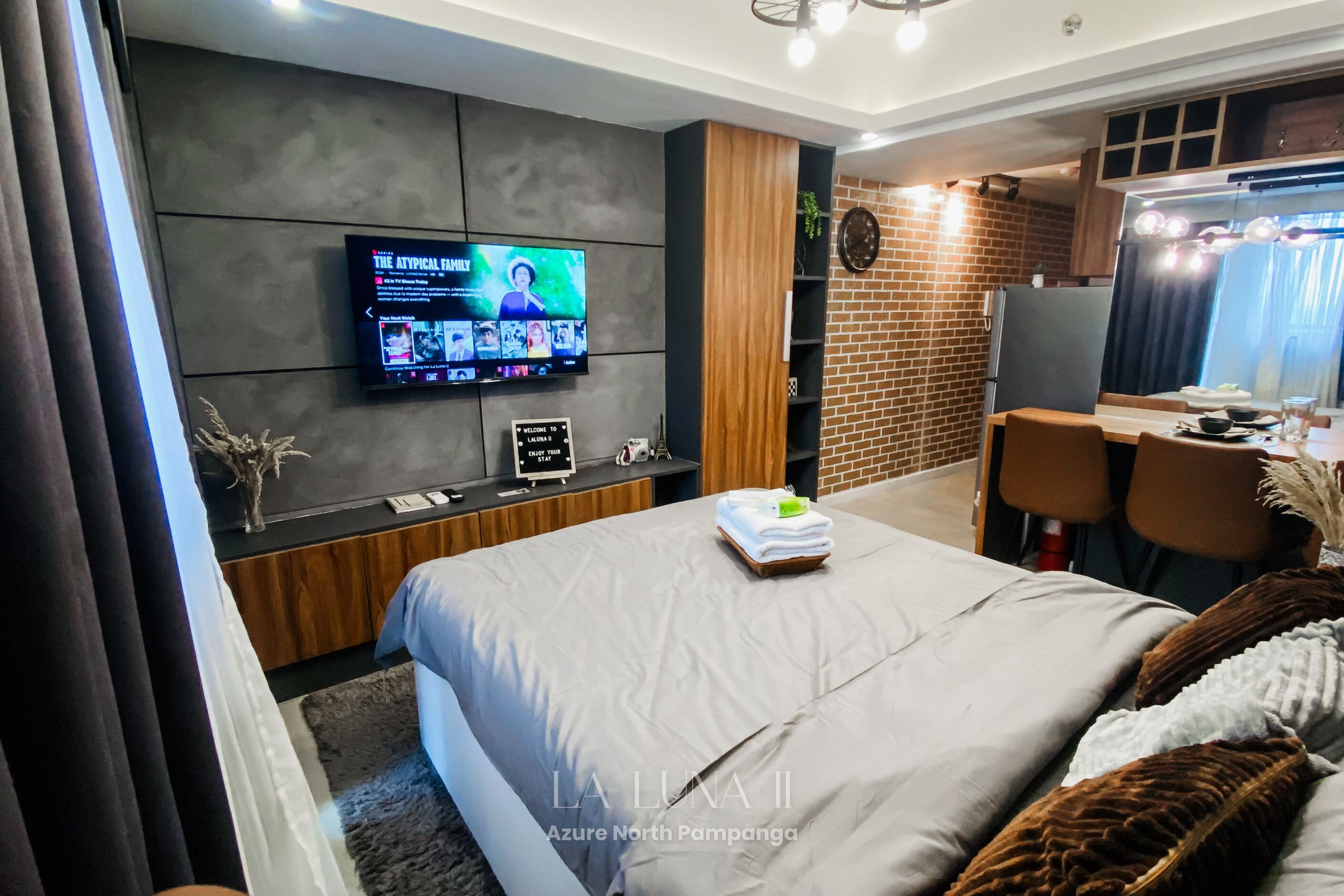 Una habitación de hotel moderna con una cama grande, un televisor de pantalla plana montado en la pared, un escritorio con una silla y un detalle de pared de ladrillo.