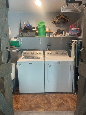 A laundry room with two front-loading washers and dryers, a shelf with various items, and a hanging clothes rack.