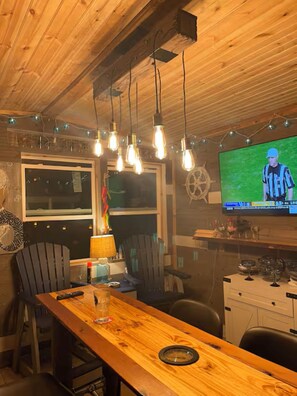 A wooden interior with a bar area, chairs, a television displaying a sports game, and a ship's wheel decoration.