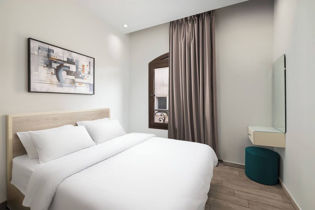 Habitación de hotel con cama, un cuadro, ventana con cortinas, un espejo y un pequeño taburete.