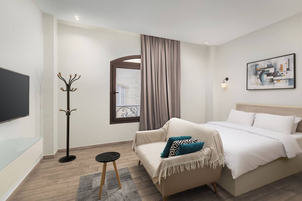 Una habitación de hotel moderna con cama, sofá, perchero, televisión de pantalla plana y ventana con cortinas.