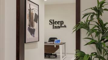 Zona de recepción de un hotel con un mostrador, una silla, una obra de arte enmarcada, un cartel que dice 'Sleep Inn' y una planta en maceta.