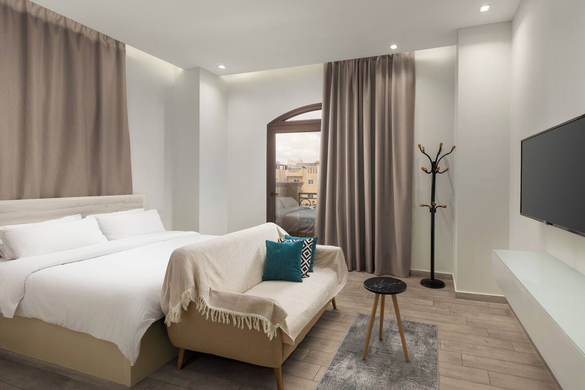 Habitación de hotel con cama, sofá, televisión, perchero y vistas a edificios.