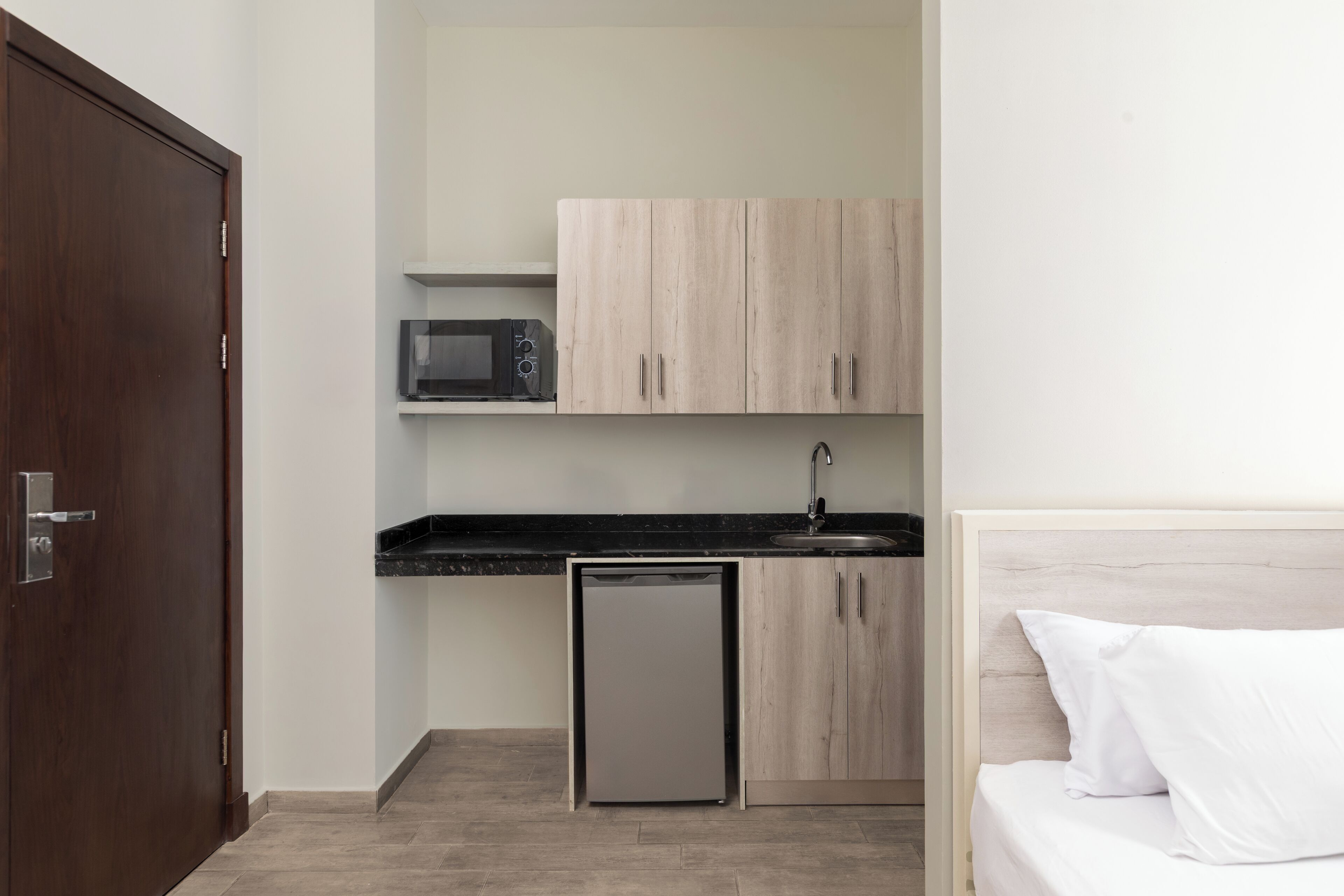 A compact room with a kitchenette, a small refrigerator, a microwave, and a bed with white bedding.
