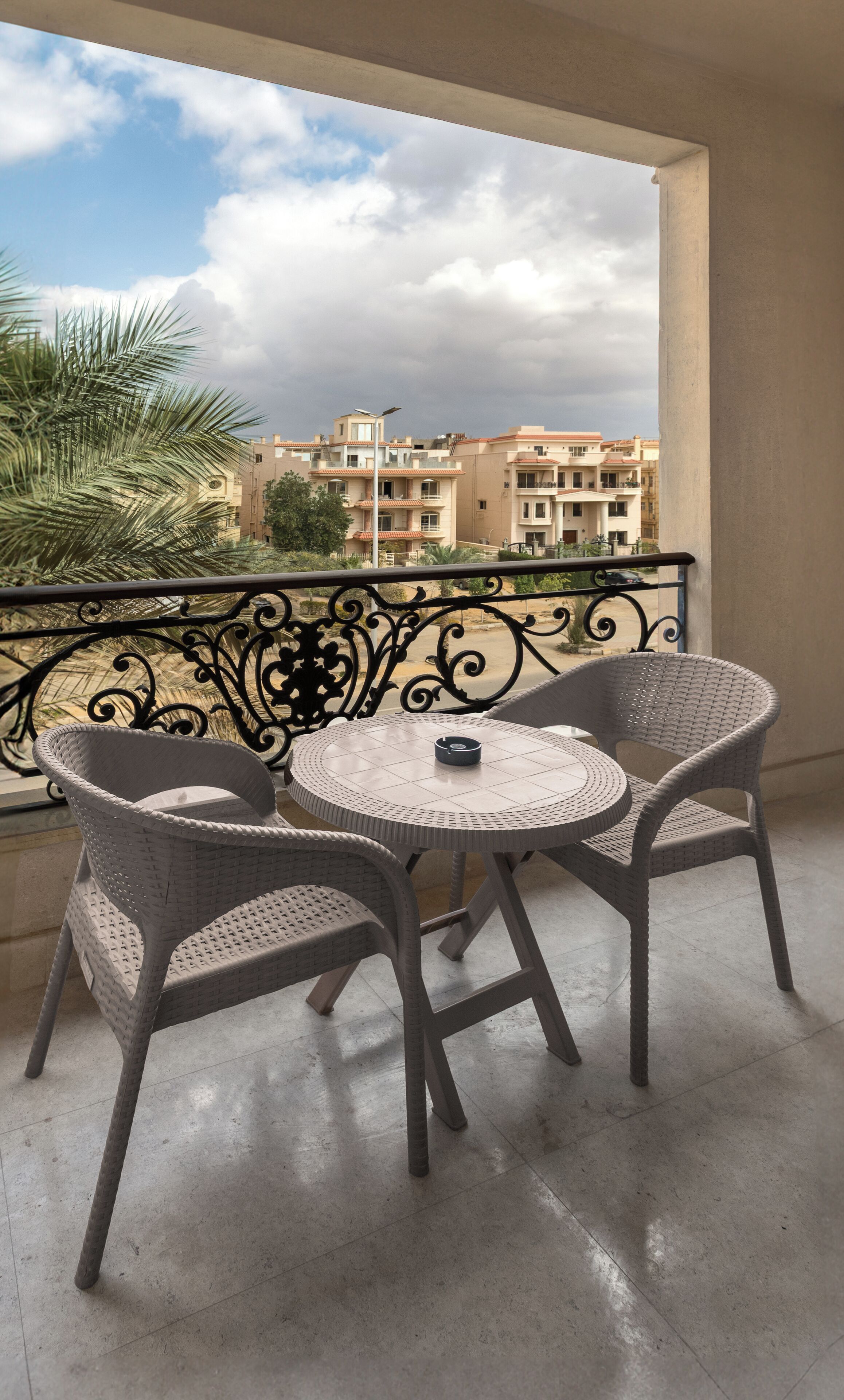 A balcony with two wicker chairs and a small round table, overlooking a residential area with buildings and a palm tree.