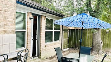 A patio with a table and chairs, a blue patterned umbrella, and a brick house with green shutters.