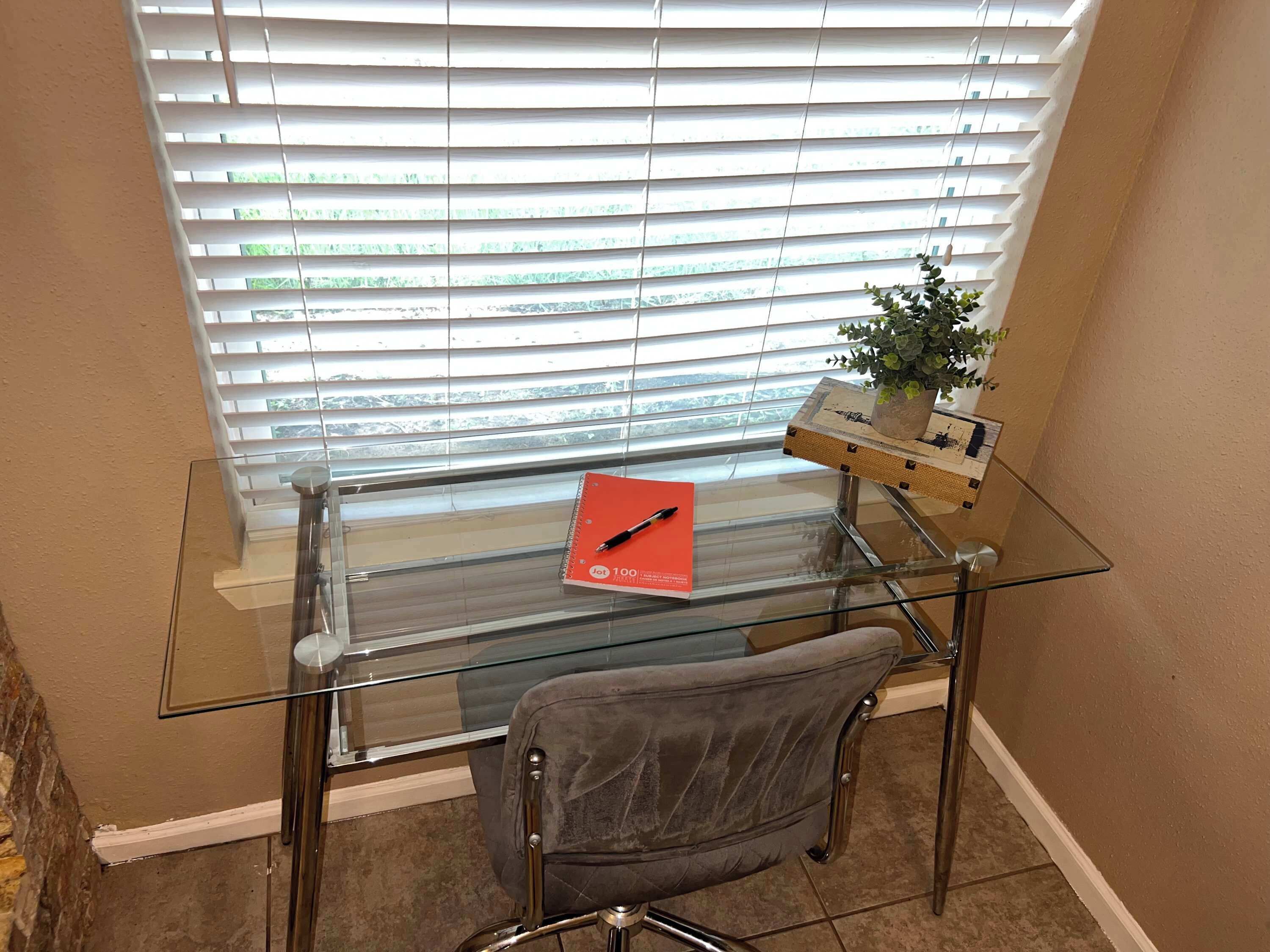 A glass desk with a red notebook and pen, a chair, and a small plant.