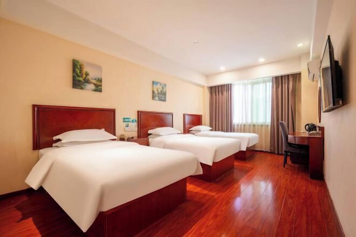 A hotel room with three beds, wooden flooring, a desk, and a television on the wall.
