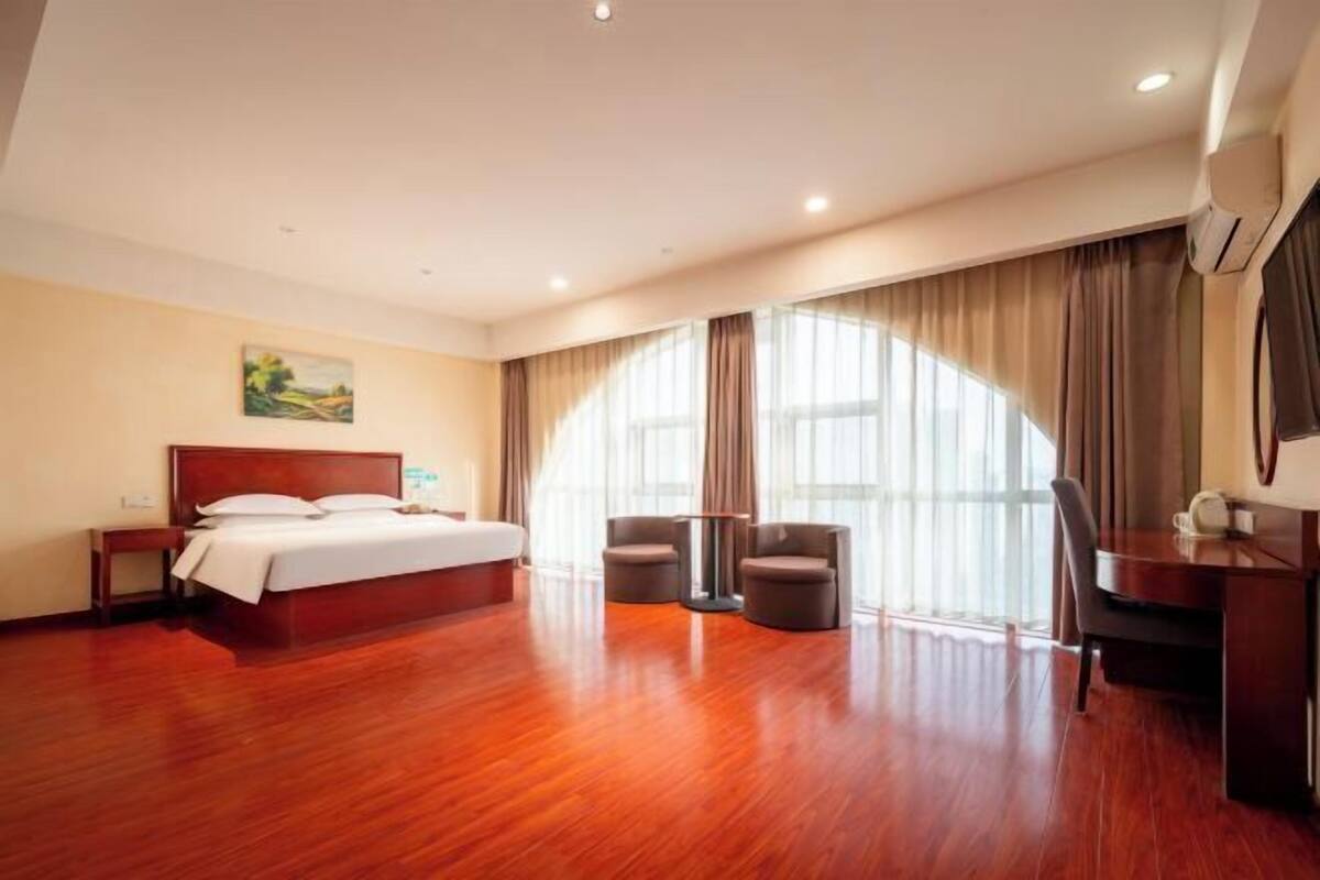 A hotel room with a wooden floor, a bed, two chairs, a desk, and a large window with curtains.
