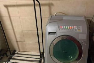 A front-loading washing machine with a control panel, a metal rack, and a beige tiled wall.